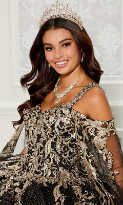 A woman smiles seated against a patterned white background, wearing the Quinceanera Dress PR30117 By Princesa—an ornate black and gold off-shoulder gown with jeweled tiara, necklace, and detachable cape.
