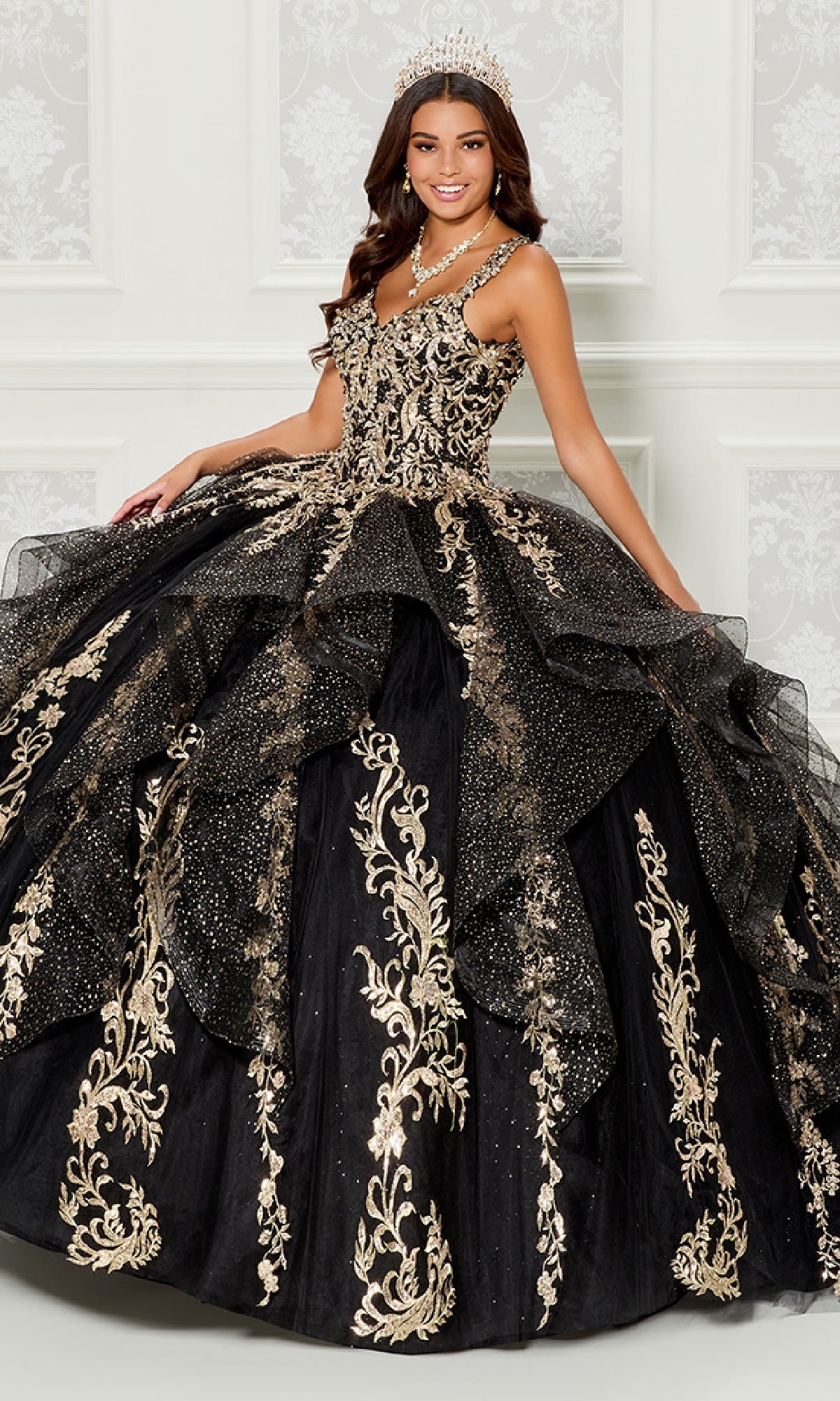 A young woman wears the Quinceanera Dress PR30117 By Princesa—an ornate ruffled tulle ball gown with gold details, full skirt, tiara, necklace, and a detachable cape—posing before a white decorative wall.