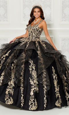 A young woman wears the Quinceanera Dress PR30117 By Princesa—an ornate ruffled tulle ball gown with gold details, full skirt, tiara, necklace, and a detachable cape—posing before a white decorative wall.