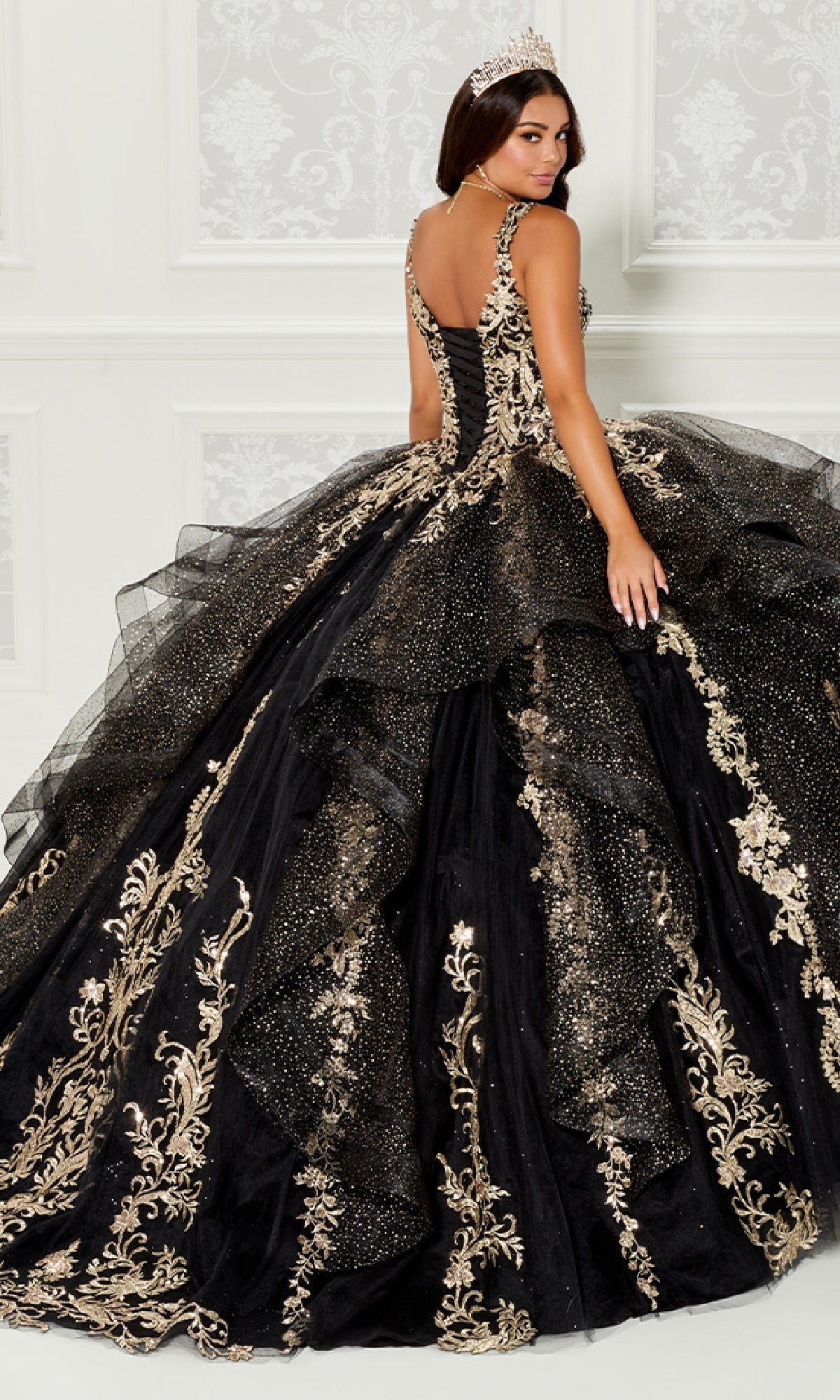 A woman wearing a tiara and the Quinceanera Dress PR30117 By Princesa, featuring ornate black fabric with gold embroidery and glitter, stands in front of a white decorative wall.
