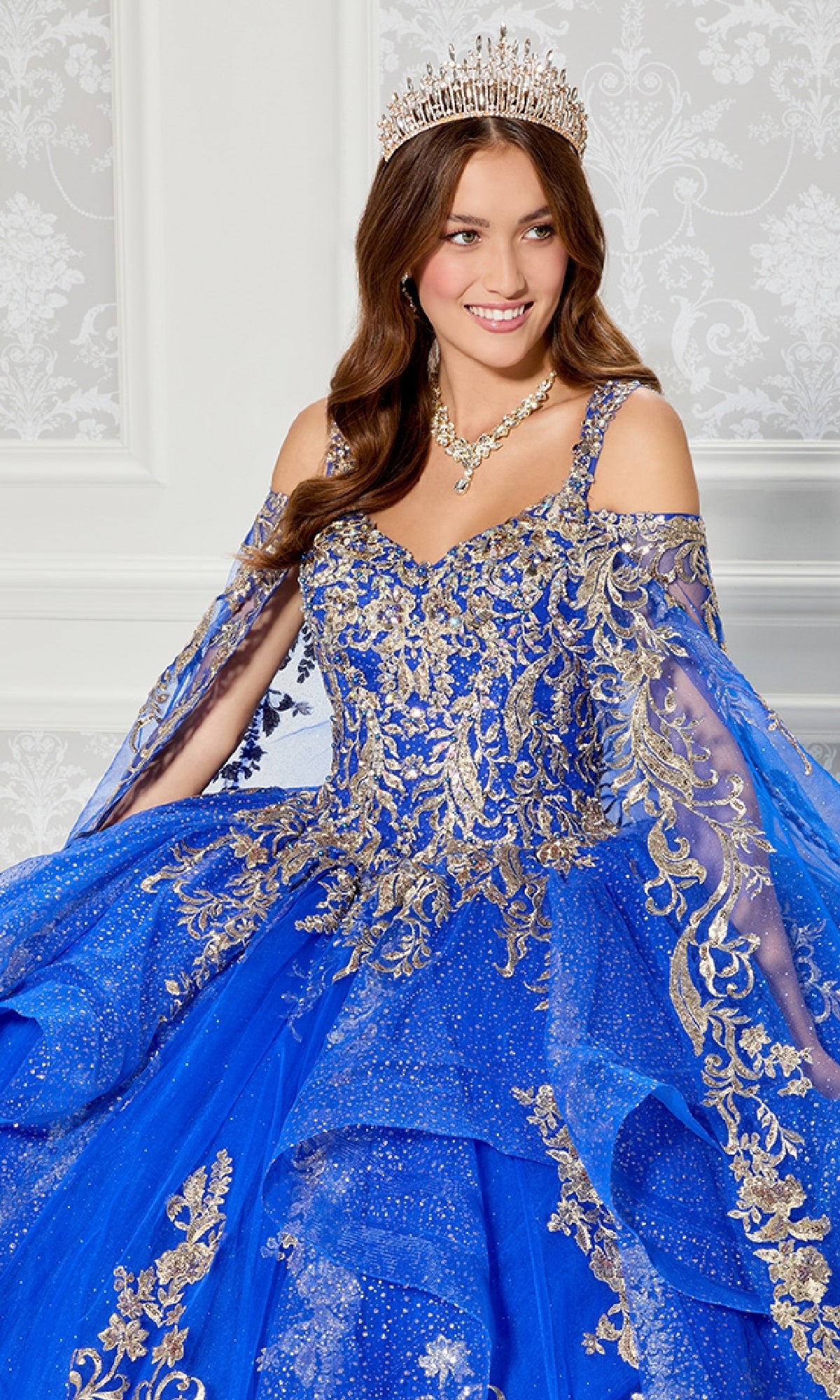 A woman stands before a patterned white wall in the Quinceanera Dress PR30117 By Princesa, featuring sparkling blue and gold details, off-the-shoulder sleeves, dramatic detachable cape, tiara, and matching jewelry.