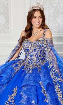 A woman stands before a patterned white wall in the Quinceanera Dress PR30117 By Princesa, featuring sparkling blue and gold details, off-the-shoulder sleeves, dramatic detachable cape, tiara, and matching jewelry.