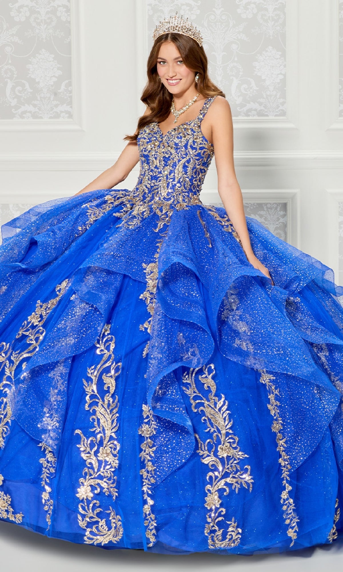 A young woman models the Quinceanera Dress PR30117 By Princesa—a sparkling royal blue gown with gold embroidery, a detachable cape, tiara, and jewelry—standing indoors against an ornate white patterned wall.