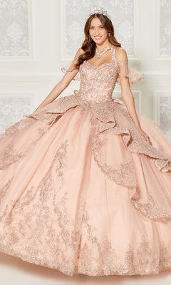 A woman in the Quinceanera Dress PR30118 By Princesa, featuring intricate lace, sequin details, and glitter embroidery, stands before a white and gray patterned wall, accessorized with a tiara and necklace.