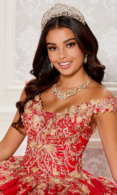 A young woman poses in front of a patterned white wall, wearing the Quinceanera Dress PR30119 By Princesa—an elegant red and gold embroidered off-the-shoulder gown with a jeweled tiara and necklace.