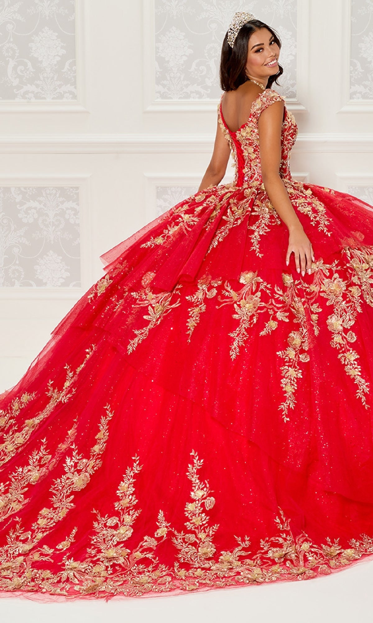 A young woman in the Quinceanera Dress PR30119 By Princesa, featuring gold embroidery, poses with a tiara in an elegant room with white walls and ornate patterns.