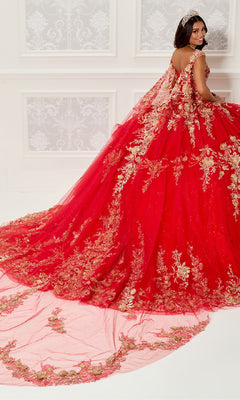 A woman wears the Quinceanera Dress PR30119 By Princesa, featuring gold embroidery and a glitter tulle gown with a long train, standing in an ornate room as her detachable cathedral cape flows elegantly behind her.