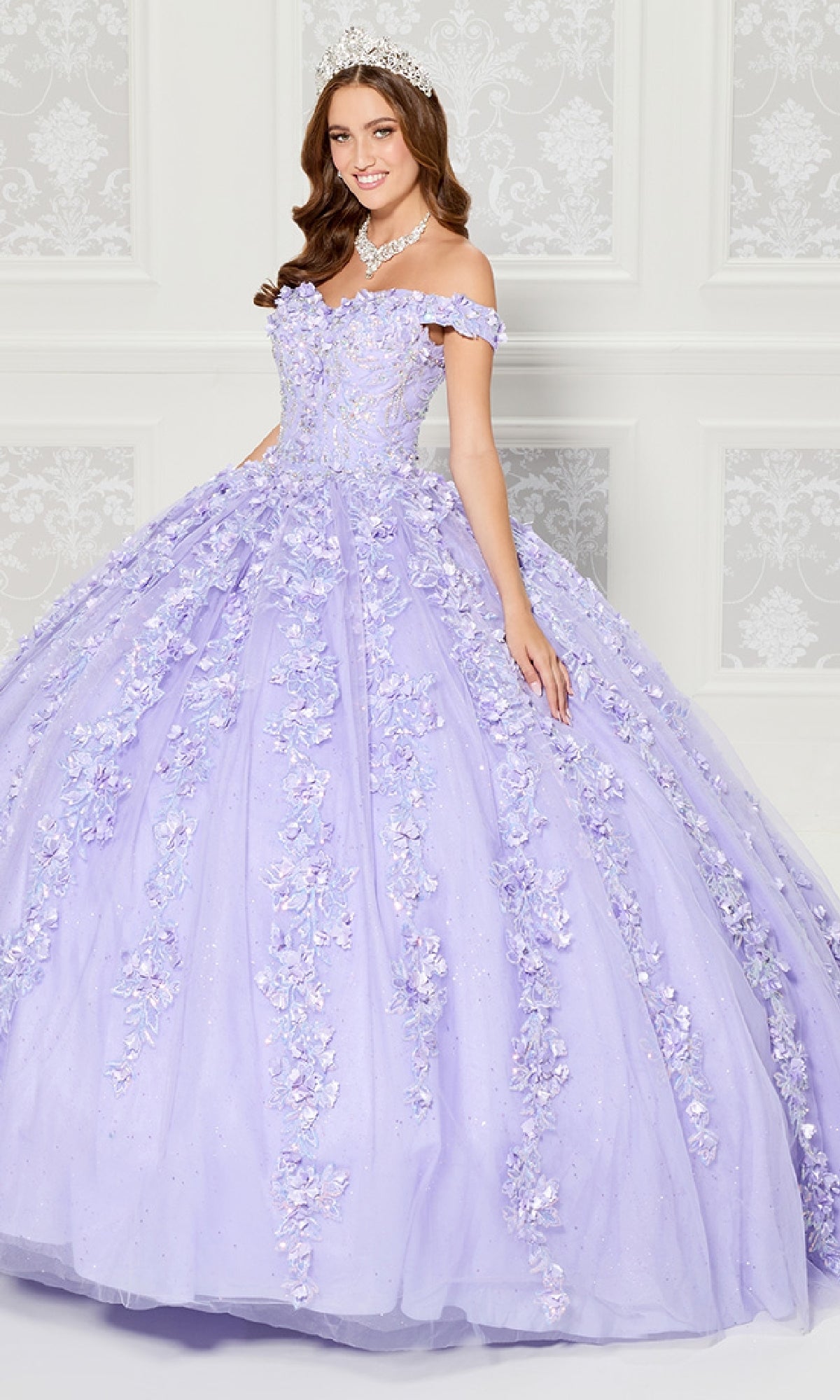 A woman in the Princess Quinceanera Dress: Princesa PR30120, featuring lavender floral tulle, 3D flowers, and an off-the-shoulder neckline, poses with a tiara and jeweled necklace before a white patterned wall.