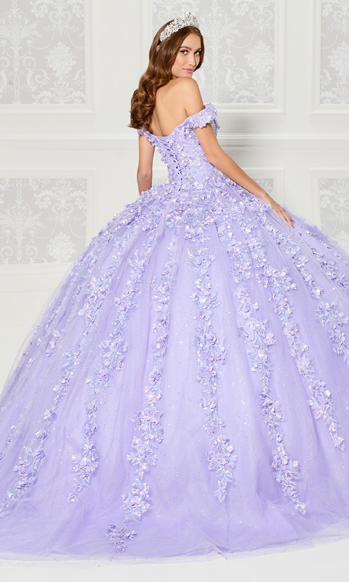 A woman wearing the Princesa PR30120 light purple off-the-shoulder quinceanera dress with 3D floral lace and a tiara stands in front of an ornate white wall, facing slightly away.
