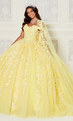 A young woman in the Princesa PR30120 Princess Quinceanera Dress, featuring jeweled tiara and yellow 3D flower-adorned ball gown, poses before a white ornate wall.