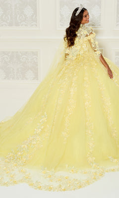 A woman in the Princess Quinceanera Dress: Princesa PR30120, a yellow floral embroidered gown with a long train, stands before a white patterned wall, glancing over her shoulder.