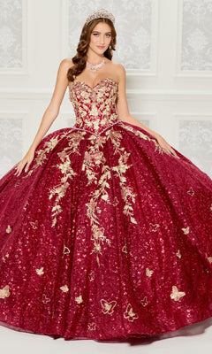 A woman in the Strapless Quinceanera Dress: Princesa PR30121, with red and gold embroidery and a full skirt, stands before a white patterned wall wearing a tiara and necklace.