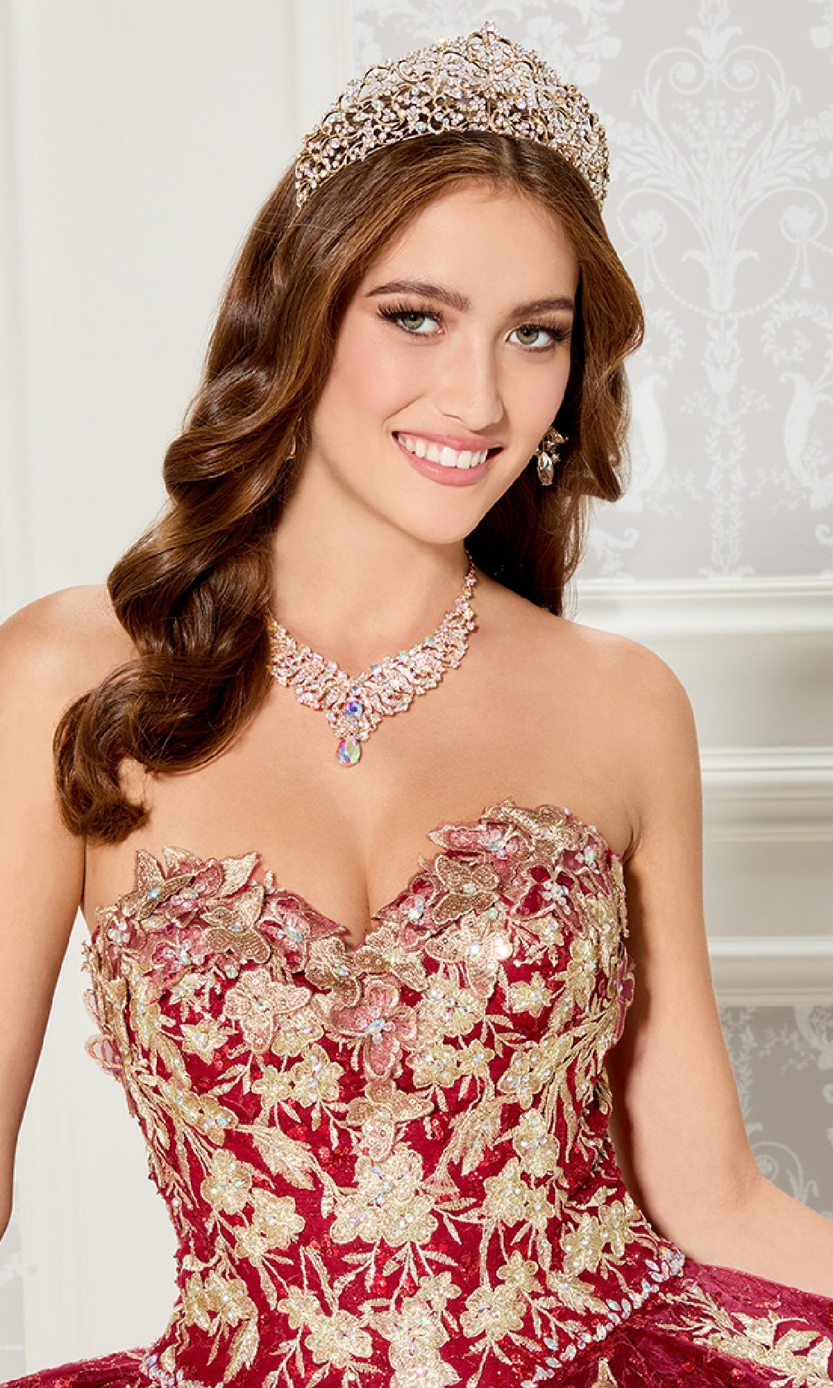 A woman in the Princesa PR30121 strapless quinceanera dress with gold floral embroidery, jeweled tiara, and drop earrings stands before a white ornate backdrop.