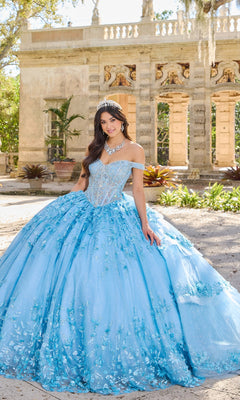 A young woman wears the Princesa PR30131 Off-Shoulder Quinceanera Dress with 3D floral appliques and a corset bodice, standing outdoors before ornate stone arches, accessorized with a tiara and necklace.