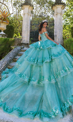 A young woman poses on stone steps amidst greenery and columns, wearing the sparkling Off-Shoulder Quinceanera Dress: Princesa PR30131 with a corset bodice and tiara, looking elegantly over her shoulder.