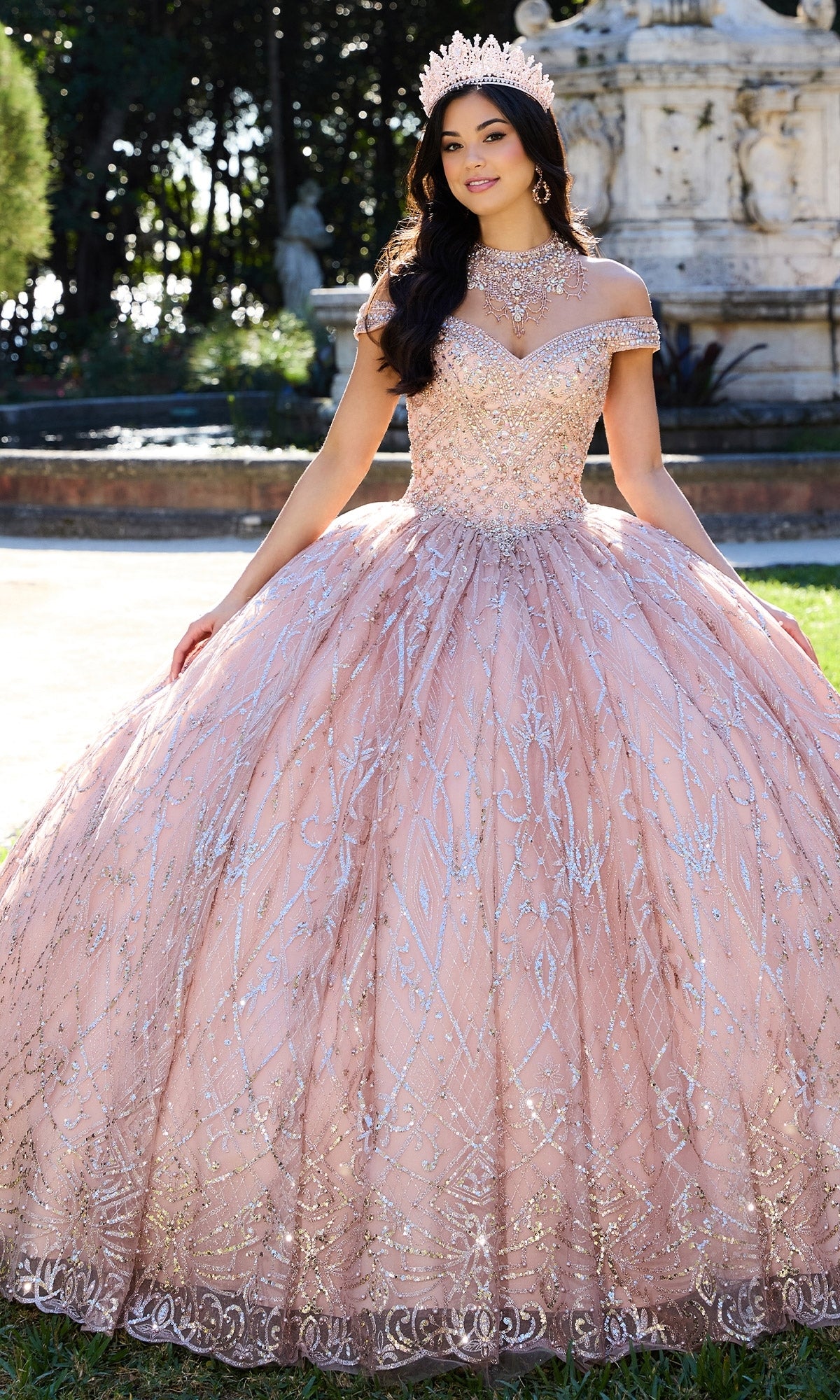 A young woman wearing the Princesa PR30132 Beaded Quinceanera Dress with silver embellishments and a crown stands outdoors before a fountain.