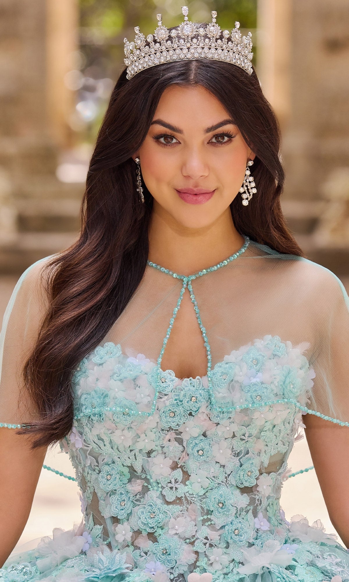 A woman stands outdoors in the Princesa PR30133 Floral Mint Quinceanera Dress, featuring 3D flowers, sheer details, a jeweled crown, and ornate earrings.
