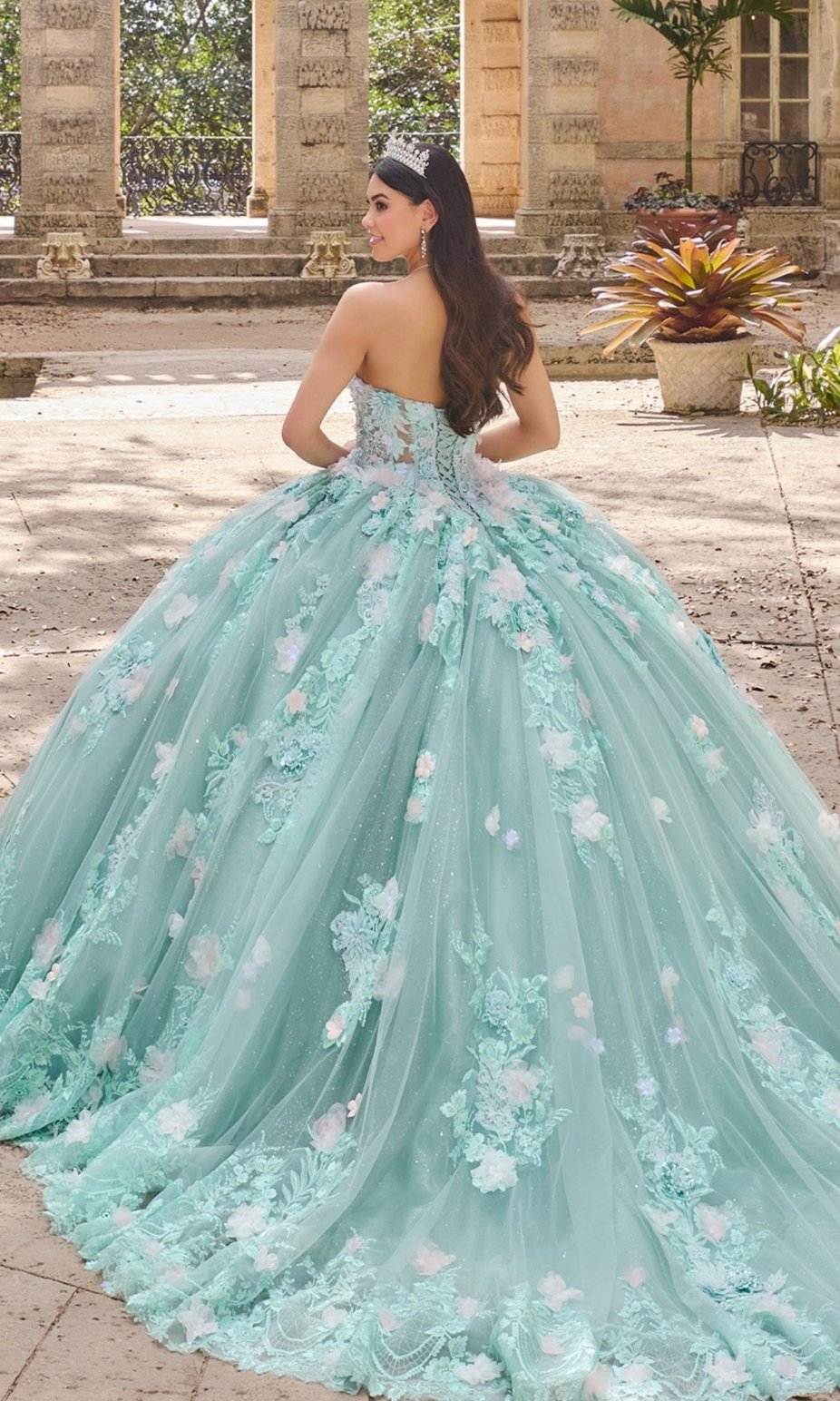 A woman stands outdoors, facing away, wearing the Floral Mint Quinceanera Dress: Princesa PR30133 with 3D flowers, in front of stone columns and a large plant.