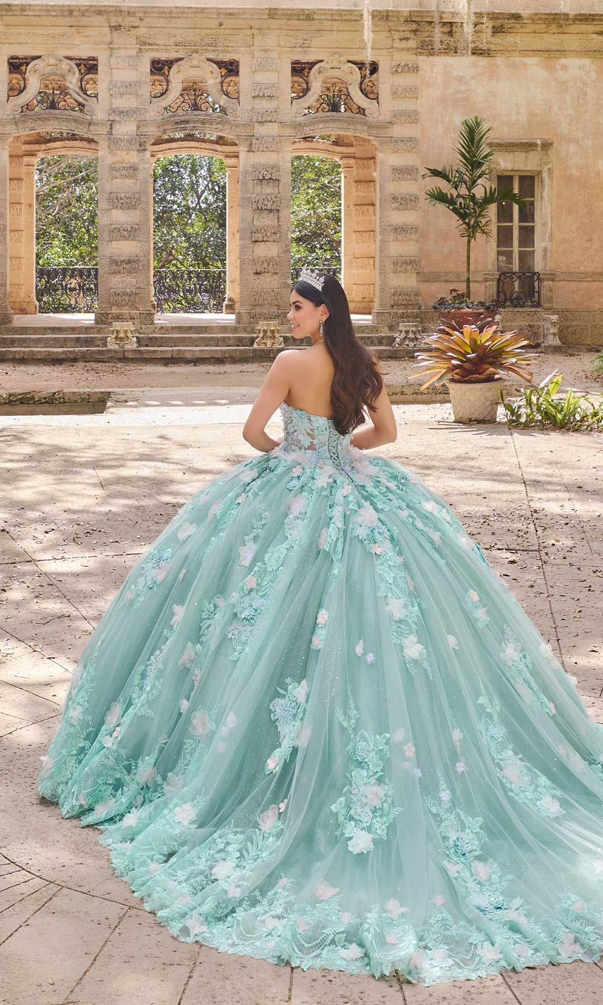 A woman in the Floral Mint Quinceanera Dress: Princesa PR30133, featuring light blue fabric and 3D flowers, stands outdoors before a historic stone structure, facing away from the camera.