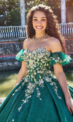 A young woman stands outdoors, smiling at the camera in the ornate Quinceanera Dress PR30134 by Princesa, featuring a green ball gown with floral embellishments and a corset bodice.