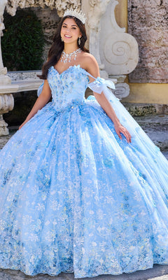 A woman stands outdoors before stone architecture wearing the 3D Floral Quinceanera Dress: Princesa PR30135, a blue gown with floral patterns, 3D flowers, off-the-shoulder sleeves, plus a jeweled necklace and tiara.