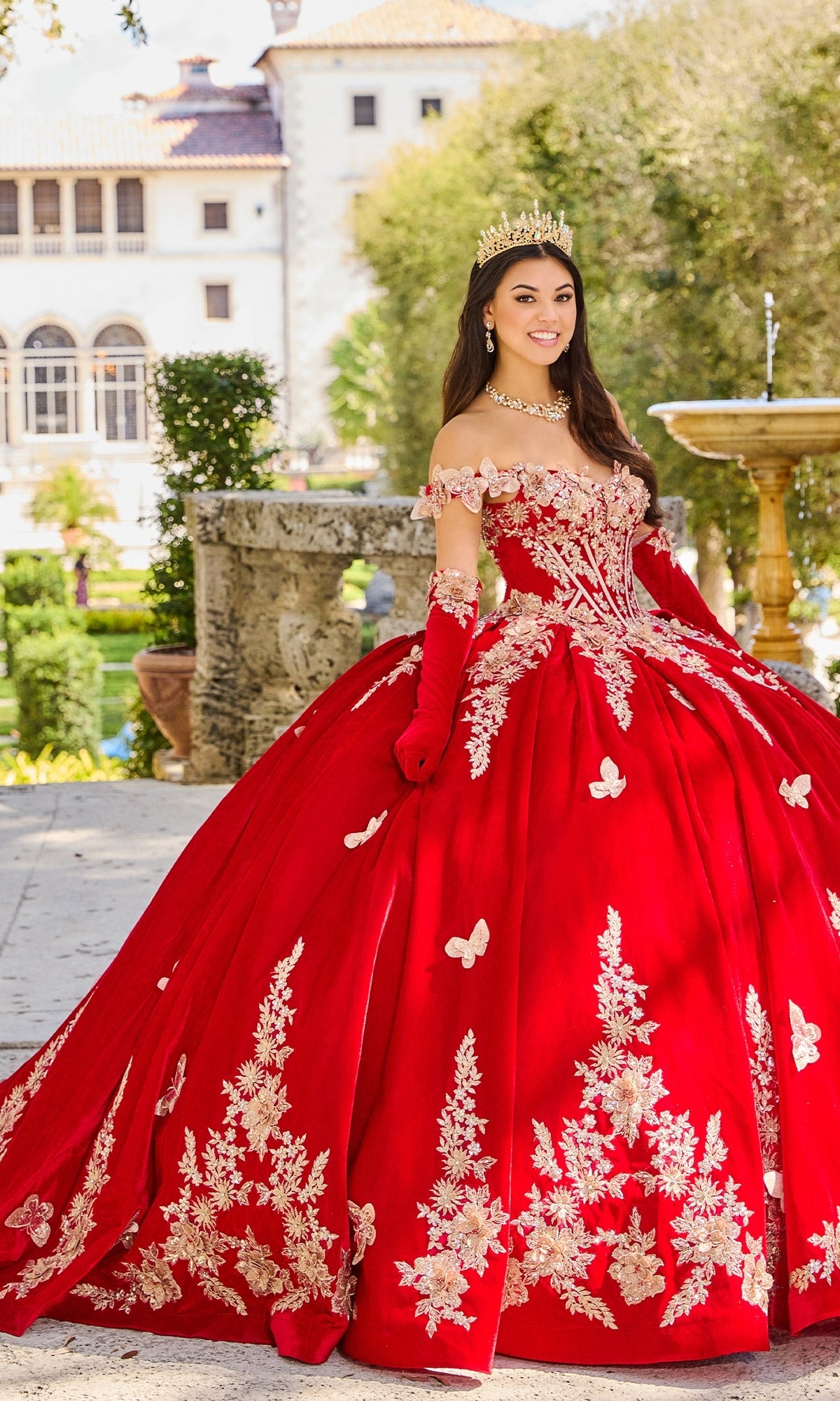 A young woman wearing the Princesa PR30136 Velvet Quinceanera Dress with gold lace embroidery and a tiara stands outdoors near a historic building and fountain.