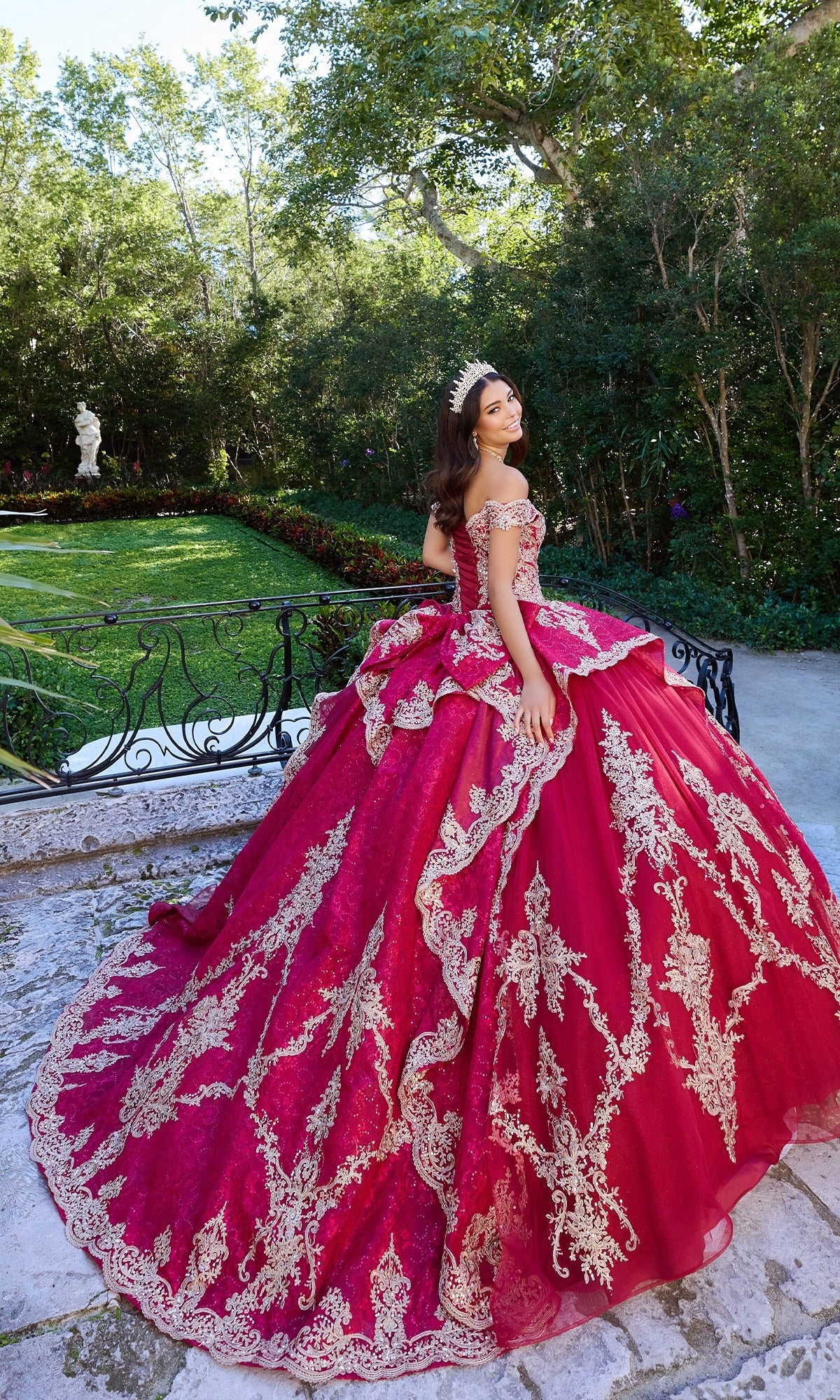A young woman stands outdoors among greenery in the Quinceanera Dress PR30138 By Princesa, a red off-the-shoulder gown with gold embroidery, sparkling glitter tulle, and a tiara, creating an elegant scene on a stone pathway.