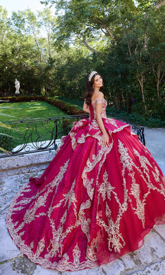 A young woman stands outdoors among greenery in the Quinceanera Dress PR30138 By Princesa, a red off-the-shoulder gown with gold embroidery, sparkling glitter tulle, and a tiara, creating an elegant scene on a stone pathway.
