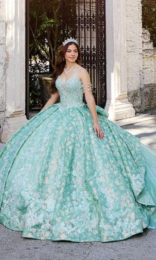 A young woman in the Quinceanera Dress PR30139 By Princesa, a light green floral gown with glitter tulle and a tiara, stands outdoors near a stone archway.