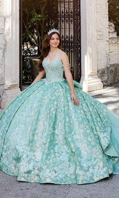 A young woman in the Quinceanera Dress PR30139 By Princesa, a light green floral gown with glitter tulle and a tiara, stands outdoors near a stone archway.