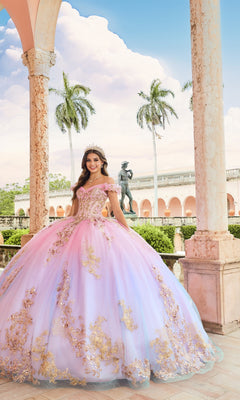 A young woman smiles under an arched stone structure, wearing the Quinceanera Dress PR30152 By Princesa with gold details and shimmering sequins, as palm trees and a statue appear in the background.