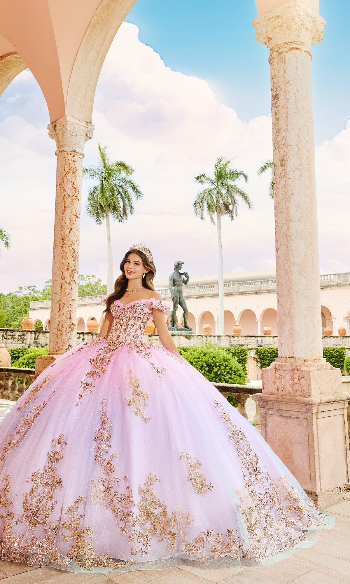 A young woman in the Quinceanera Dress PR30152 By Princesa, featuring pink and gold shimmering sequins, stands under a stone archway with palm trees and a statue in the background.