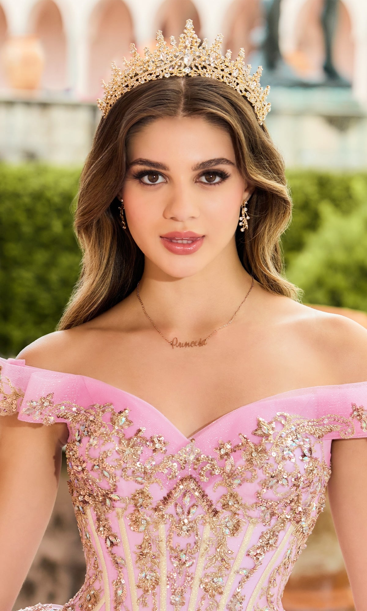 A young woman with long brown hair stands outdoors wearing the Quinceanera Dress PR30152 by Princesa, a pink off-shoulder gown with shimmering sequins, paired with a gold crown and a 