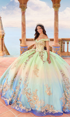 Wearing the Quinceanera Dress PR30152 By Princesa, a woman in a pastel gown with gold details and lace appliques stands on a seaside terrace with a tiara, smiling.