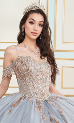 A woman wearing a jeweled crown and the Princesa PR30153 Lace Embroidered Quinceanera Dress with an ornate off-the-shoulder lace corset bodice stands before a decorative wall.