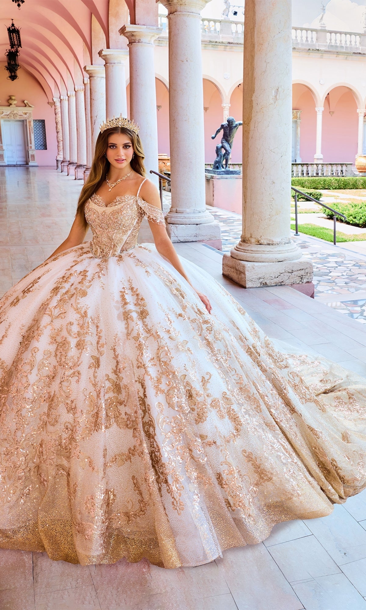 A woman wearing the Lace Embroidered Quinceanera Dress: Princesa PR30153, featuring a gold and white corset bodice and tiara, stands in a pink hallway with columns and arched ceilings.