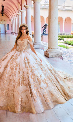 A woman wearing the Lace Embroidered Quinceanera Dress: Princesa PR30153, featuring a gold and white corset bodice and tiara, stands in a pink hallway with columns and arched ceilings.