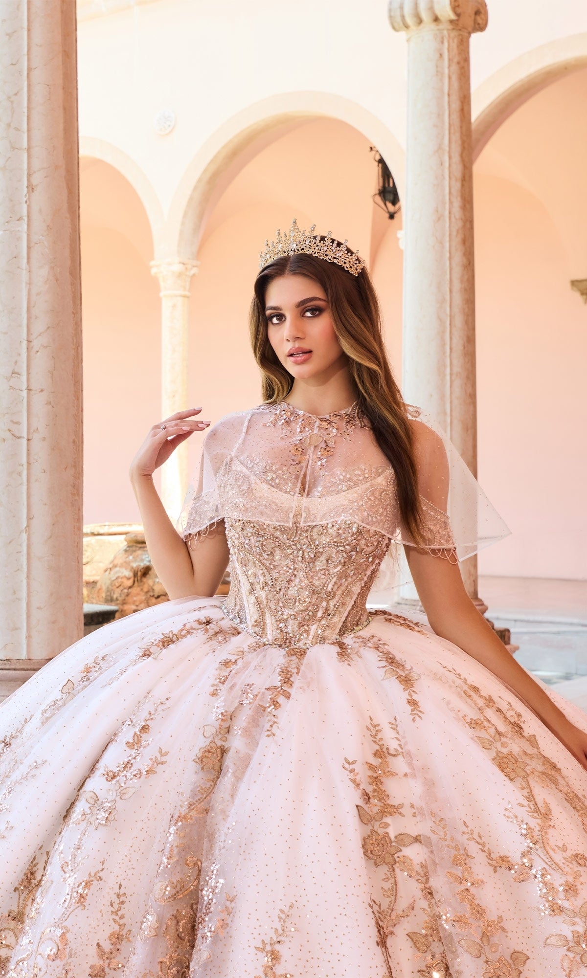 A young woman wears the Princesa PR30153 Lace Embroidered Quinceanera Dress with gold embellishments and a corset bodice, standing in a courtyard with arched columns, looking at the camera while wearing a tiara.