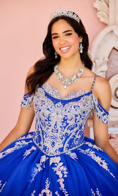A young woman in the Lace Embroidered Quinceanera Dress: Princesa PR30153, featuring a tiara, blue fabric with silver details, and a corset bodice, smiles at the camera against a pink background.