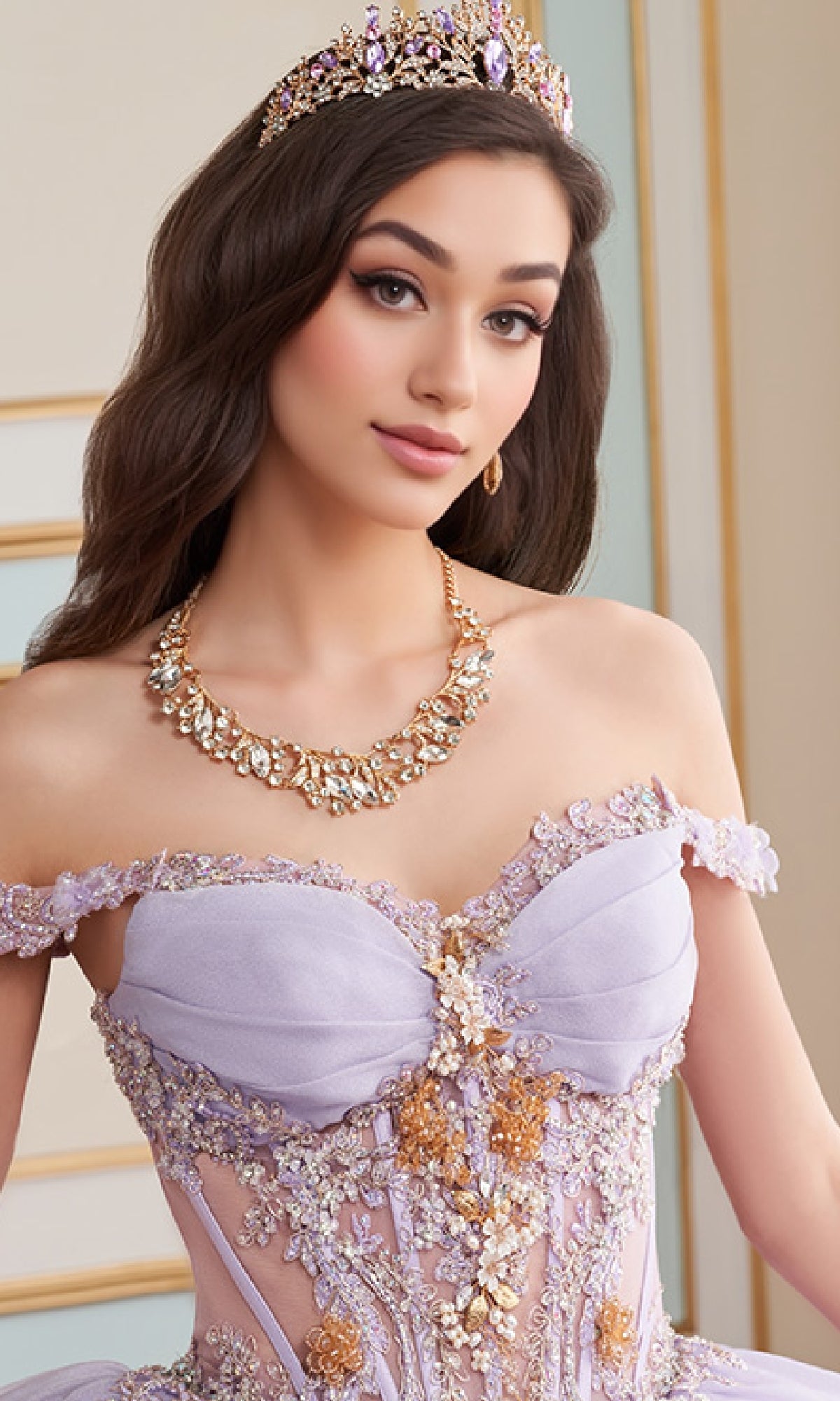 A woman wearing a jeweled crown, ornate gold necklace, and the Quinceanera Dress PR30154 By Princesa—an off-shoulder lavender ball gown with a sweetheart neckline—poses indoors.
