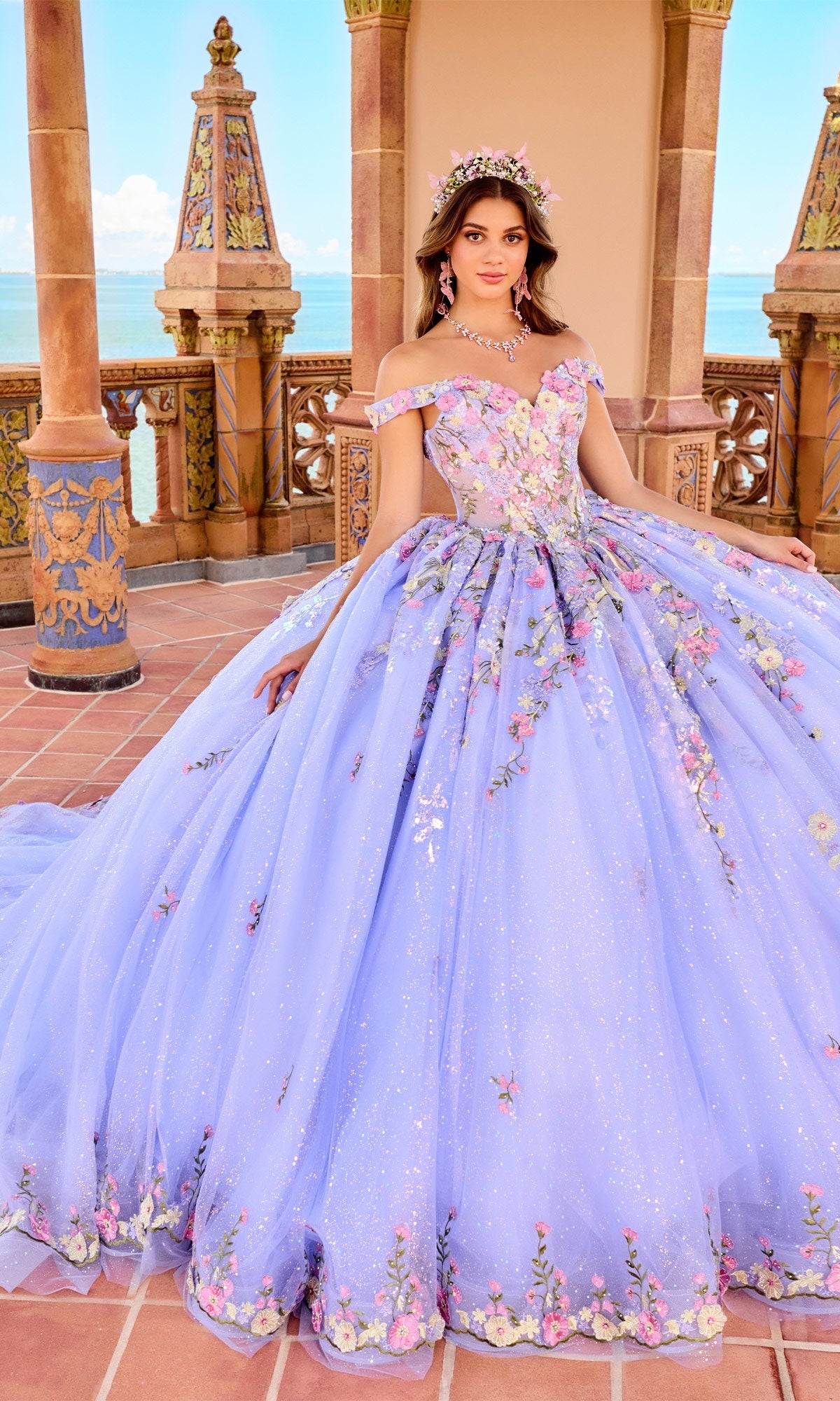 A woman in the Quinceanera Dress PR30155 By Princesa, a large off-the-shoulder lavender gown with 3D floral embroidery, stands on a terrace overlooking the ocean.