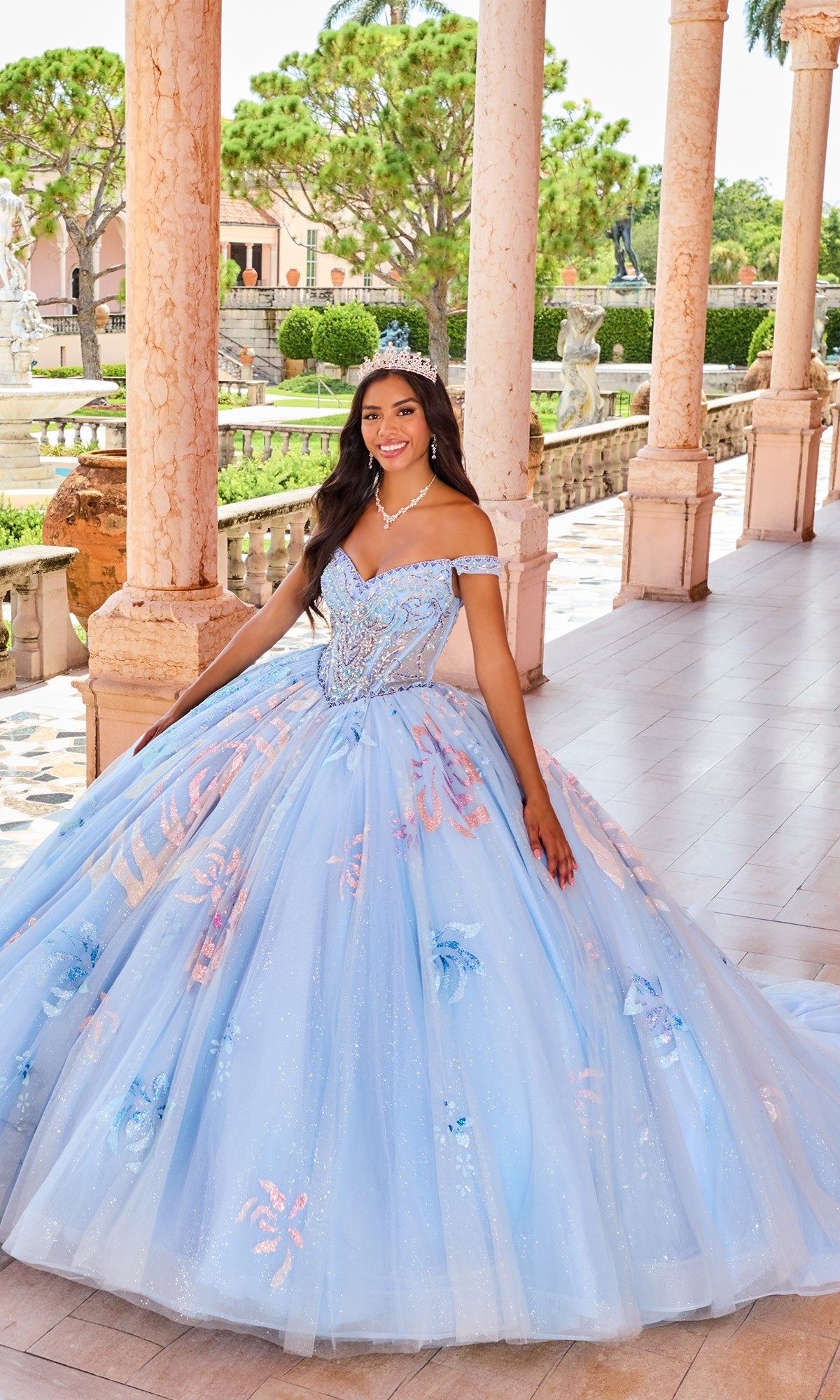 A young woman wears the Quinceanera Dress PR30156 By Princesa with detachable off-the-shoulder straps and a tiara, standing on a tiled terrace framed by columns and lush greenery.