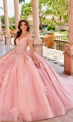A young woman in the Quinceanera Dress PR30156 By Princesa, featuring a pink hue, sweetheart neckline, and detachable off-the-shoulder straps, smiles on a sunlit stone terrace framed by columns and lush greenery.