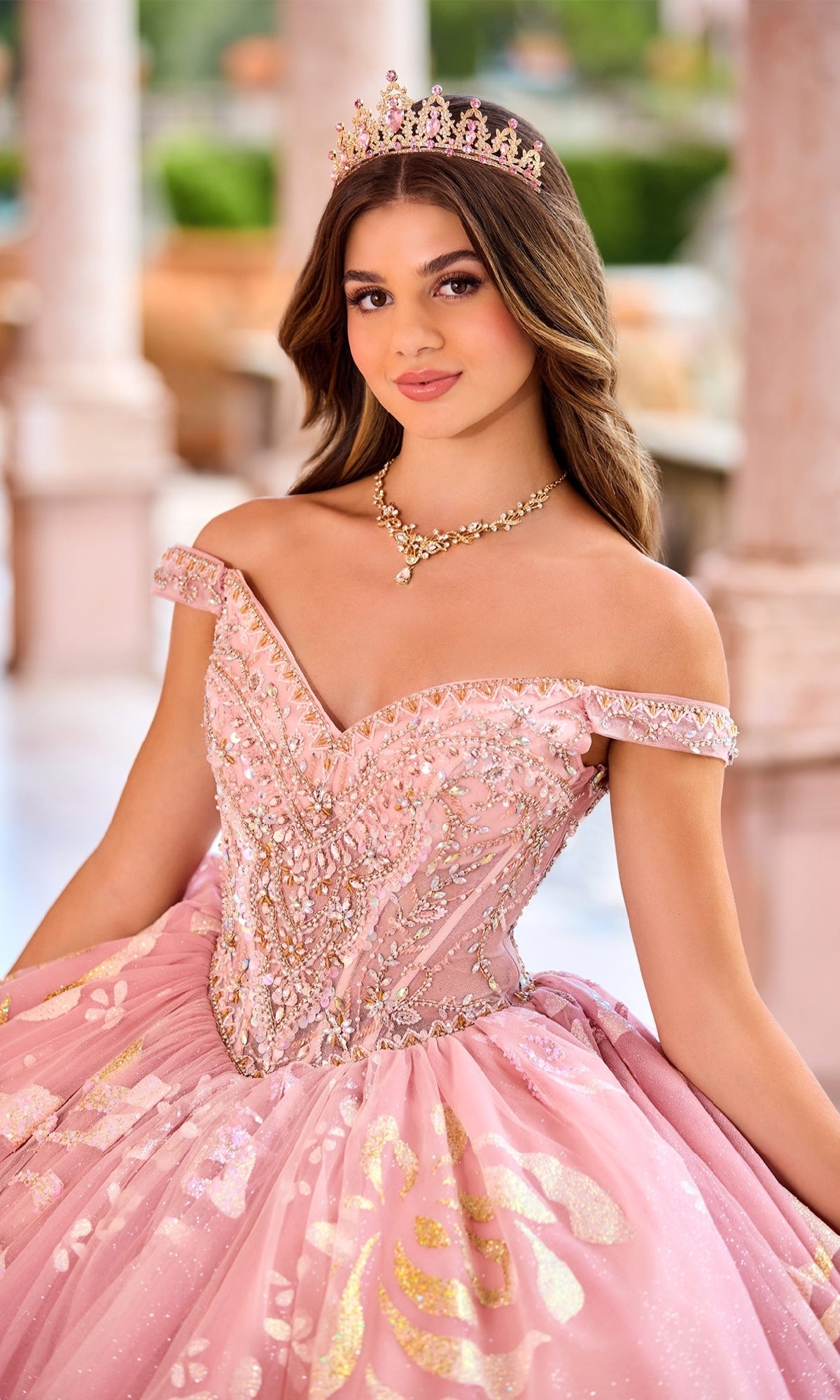 A young woman stands in a formal outdoor setting wearing the Princesa Quinceanera Dress PR30156, featuring a jeweled tiara, sweetheart neckline, ornate pink design, and detachable off-the-shoulder straps.