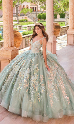 A young woman wears the Quinceanera Dress PR30157 By Princesa, featuring light blue glitter details, 3D flowers, lace appliques, and a sparkling tiara as she poses on a columned terrace in a formal garden.