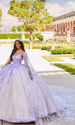 A young woman stands outdoors in a formal garden near an arched, statue-adorned building, wearing the shimmering Quinceanera Dress PR30158 By Princesa—a lavender gown with a lace cape and delicate embroidered details.