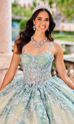 A woman in a jeweled tiara and necklace smiles outdoors, wearing the Quinceanera Dress PR30158 By Princesa—a light blue gown with sequins and floral details.