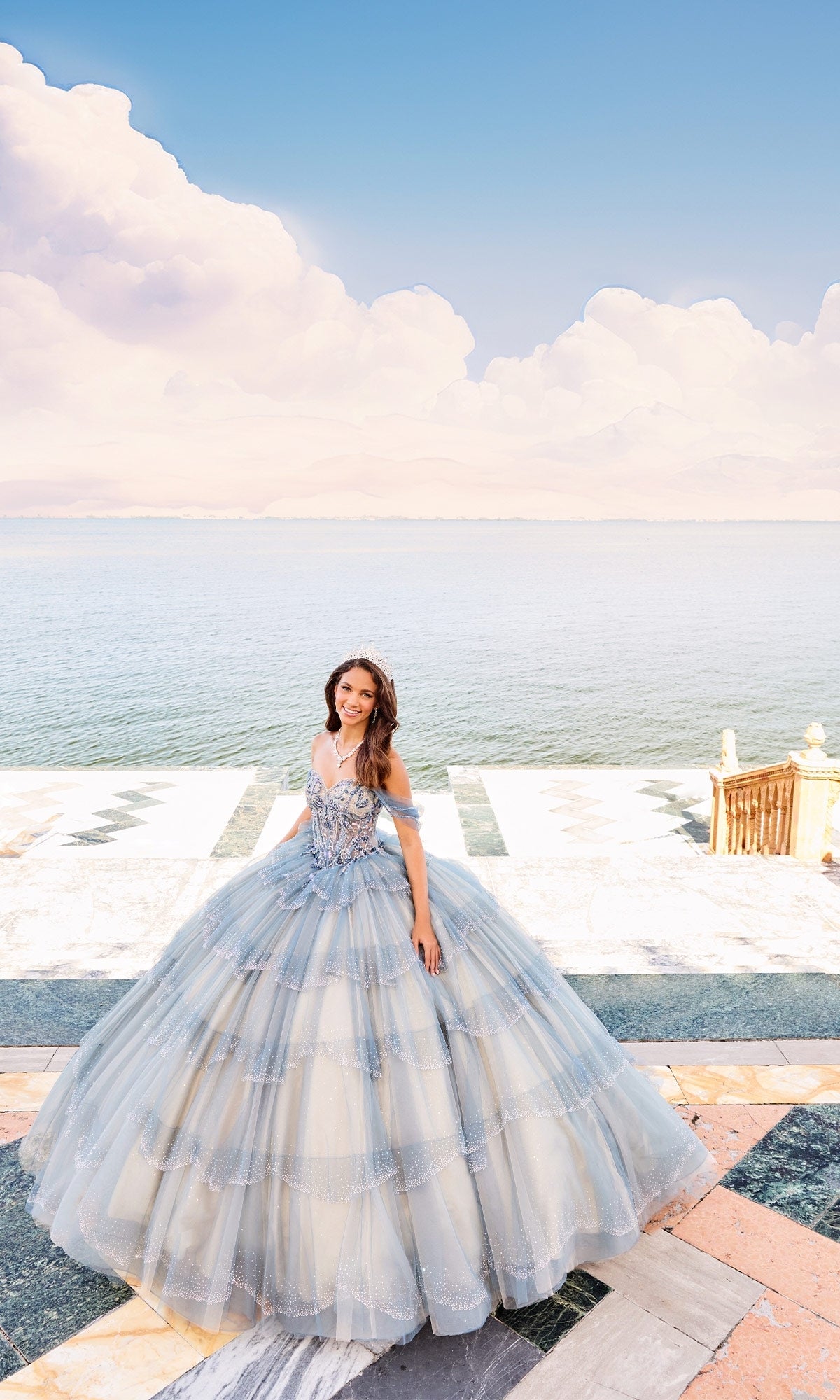 A young woman in the Quinceanera Dress PR30160 by Princesa, a light blue glitter tulle ball gown with an off-the-shoulder bodice, stands on a marble terrace overlooking tranquil water beneath a softly clouded blue sky.