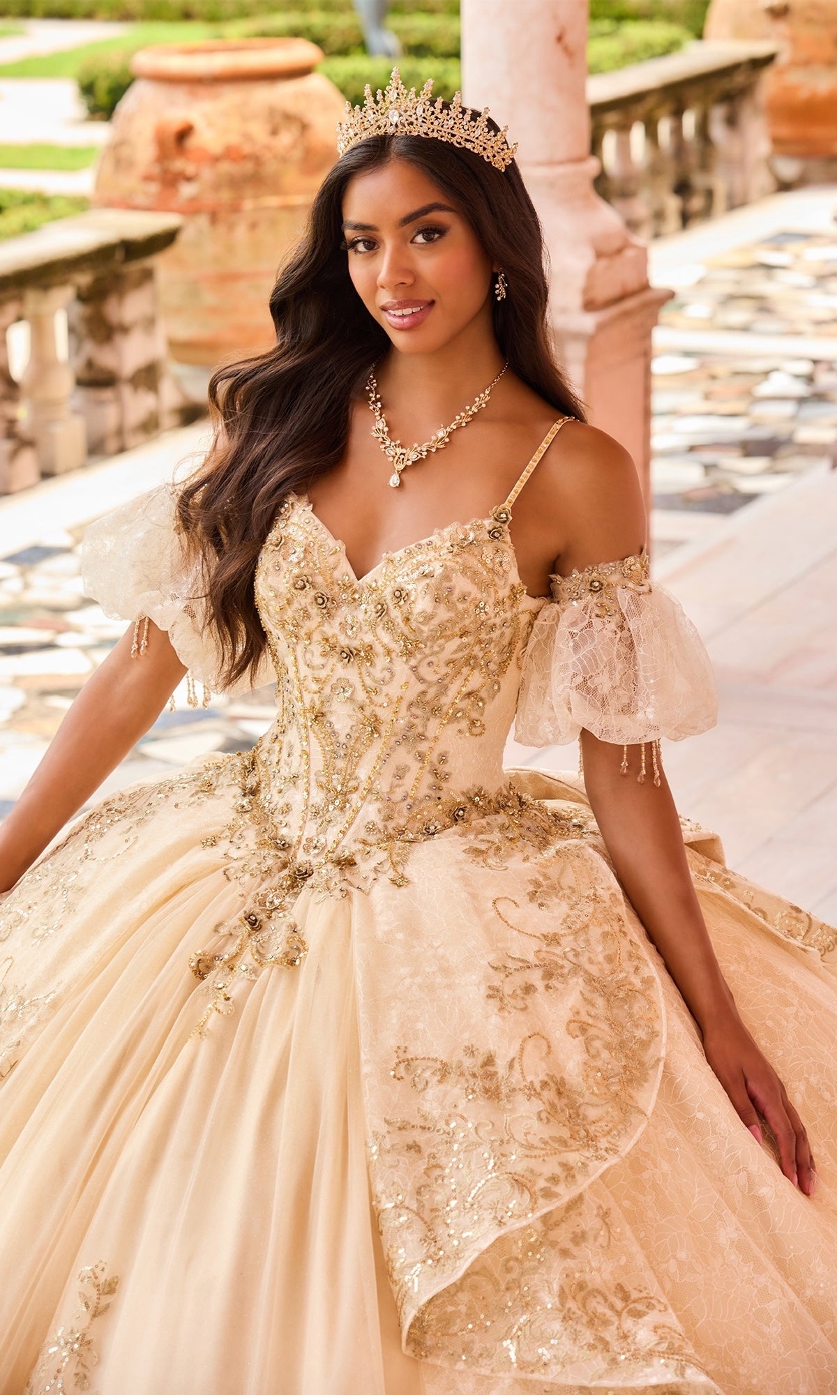 A woman in the Princesa PR30161 Lace Quinceanera Dress, adorned with gold embellishments and glitter tulle, wears a jeweled crown while standing on a marble terrace with ornate railings and greenery in the background.