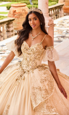 A woman in the Princesa PR30161 Lace Quinceanera Dress, adorned with gold embellishments and glitter tulle, wears a jeweled crown while standing on a marble terrace with ornate railings and greenery in the background.